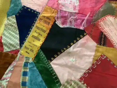 Crazy Quilt Ribbon A colorful patchwork quilt features various fabrics and stitching. One patch has gold text reading 12th Annual RE-UNION Pennsylvania Settlers Association August 30th, 1899..
