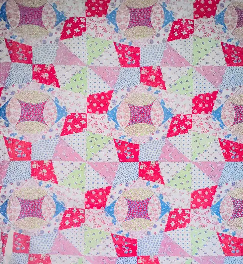A colorful patchwork quilt with geometric patterns in pink, red, blue, green, and white fabrics—this cheater cloth faux patchwork features floral and polka dot prints arranged in a repeating star and diamond design.