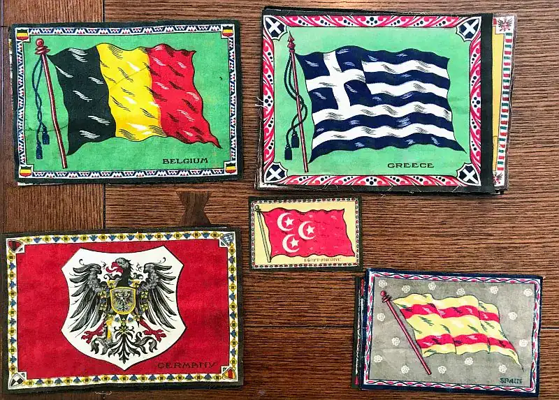 Five illustrated vintage flags labeled Belgium, Greece, Germany, Levant States, and Spain are arranged on a wooden surface, each with colorful geometric borders—a charming glimpse into the QHL Archives April 11-15 collection.