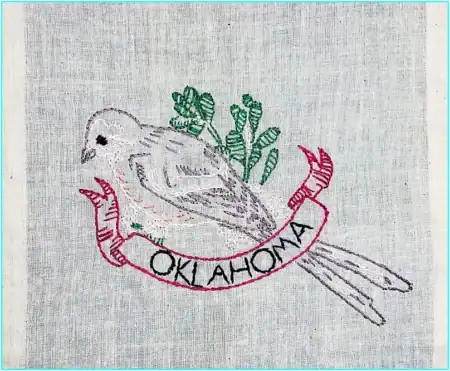 Embroidered bird with green leaves behind it, perched on a red banner that reads OKLAHOMA on light fabric—a charming example of State Birds and Flowers Outline Embroidery.