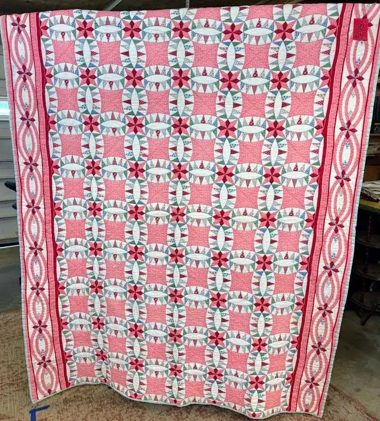A large quilt with a geometric pattern in pink, red, and white, featuring circular designs and a decorative red border with interlocking rings. The quilt is displayed vertically indoors.