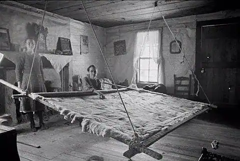 Two people work on a large, suspended wooden loom in a rustic room with wooden floors, a fireplace, and framed pictures on the mantel. Light streams in through a window.