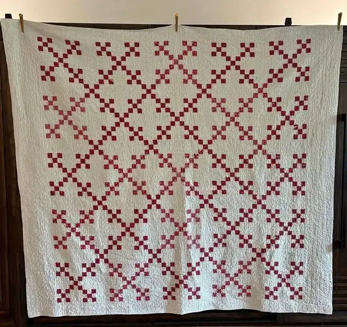 A handmade quilt with a white background features a repeating geometric pattern of red squares arranged in X and cross shapes, creating a lattice effect across the entire quilt. The quilt hangs flat against a wall.