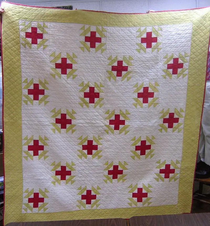 red-cross-crown A handmade quilt featuring a repeating red cross and yellow triangle pattern on a white background, bordered by a wide yellow band. The quilt is displayed vertically indoors.