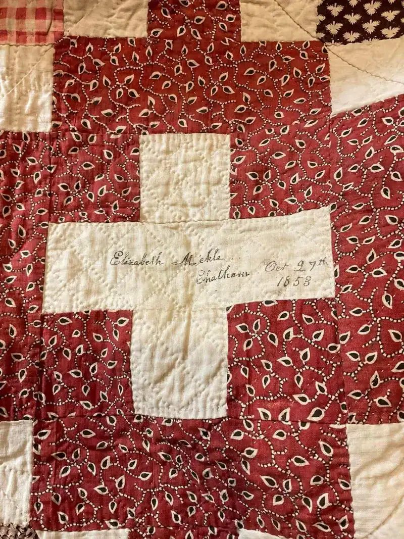 A quilt with a red and white cross pattern. The white cross has handwritten text: Elizabeth Micheli, Chatham, Oct 27th, 1888. The surrounding fabric has a red background with a small white and black paisley design.