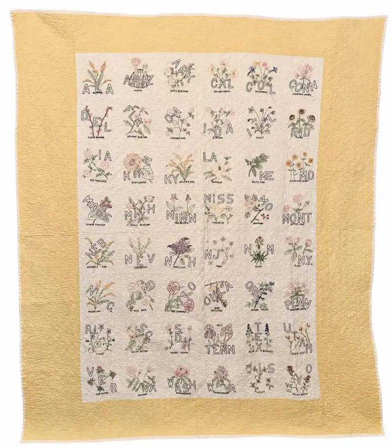 state-bird-quilt-rainbow A quilt with embroidered state abbreviations and flowers for all 50 U.S. states, arranged in a grid on a cream background with a yellow border.