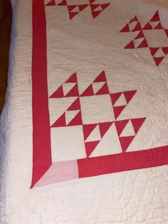 A handmade quilt with a pink and white pattern featuring geometric triangle designs and a pink border, shown on top of a bed.