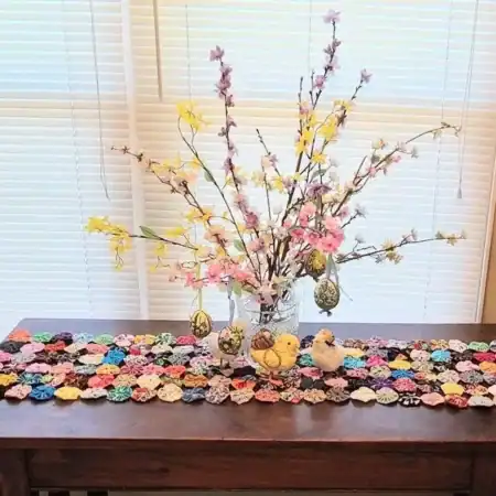 table-runner-by-yo-yo-cottage A table with a colorful quilted runner, two small bird figurines, decorated eggs, and a vase with blooming branches in front of white window blinds.