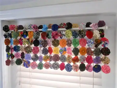 valance-by-yo-yo-cottage A row of colorful, round fabric yo-yos strung together forms a valance over a window with white blinds. The yo-yos are arranged in a grid pattern and hang from a white rod.