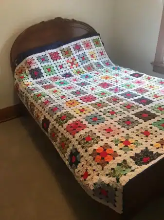 yo-yo-sewn-to-blue-back A wooden bed with a colorful, handmade patchwork quilt featuring various geometric and floral patterns, set in a simple room with beige carpet and light walls.