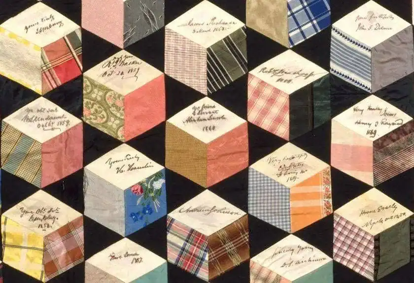 1856-tumbling-blocks-quilt A patchwork quilt featuring cube-like patterns from various colorful fabrics, each piece with handwritten names and dates, likely commemorating individuals or events.