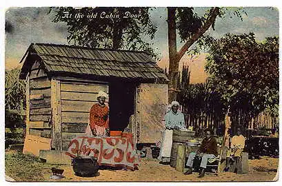 A vintage postcard shows four Black people outside a small wooden cabin, with laundry and household items nearby. The text reads, At the old Cabin Door. Trees and a fence are in the background.
