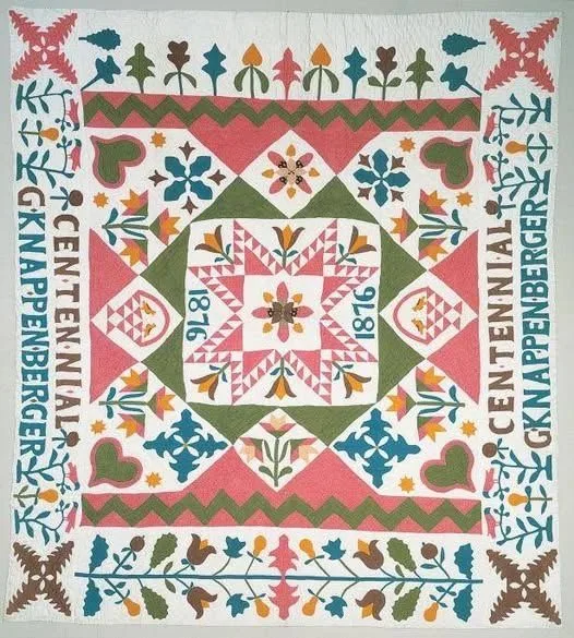 Colorful quilt with geometric and floral patterns, crafted from authentic 1876 Centennial Fabric and featuring the text CENTENNIAL! G. KNAPPENBERGER 1876 along the edges in pink. Central star design and symmetrical motifs in green, pink, blue, and gold on white.