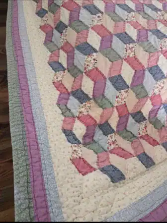 A handmade quilt with a geometric pattern featuring pink, blue, cream, and floral fabrics. It has a patchwork design and is bordered by pale blue, green, and lavender strips. The quilt is draped over a wooden surface.