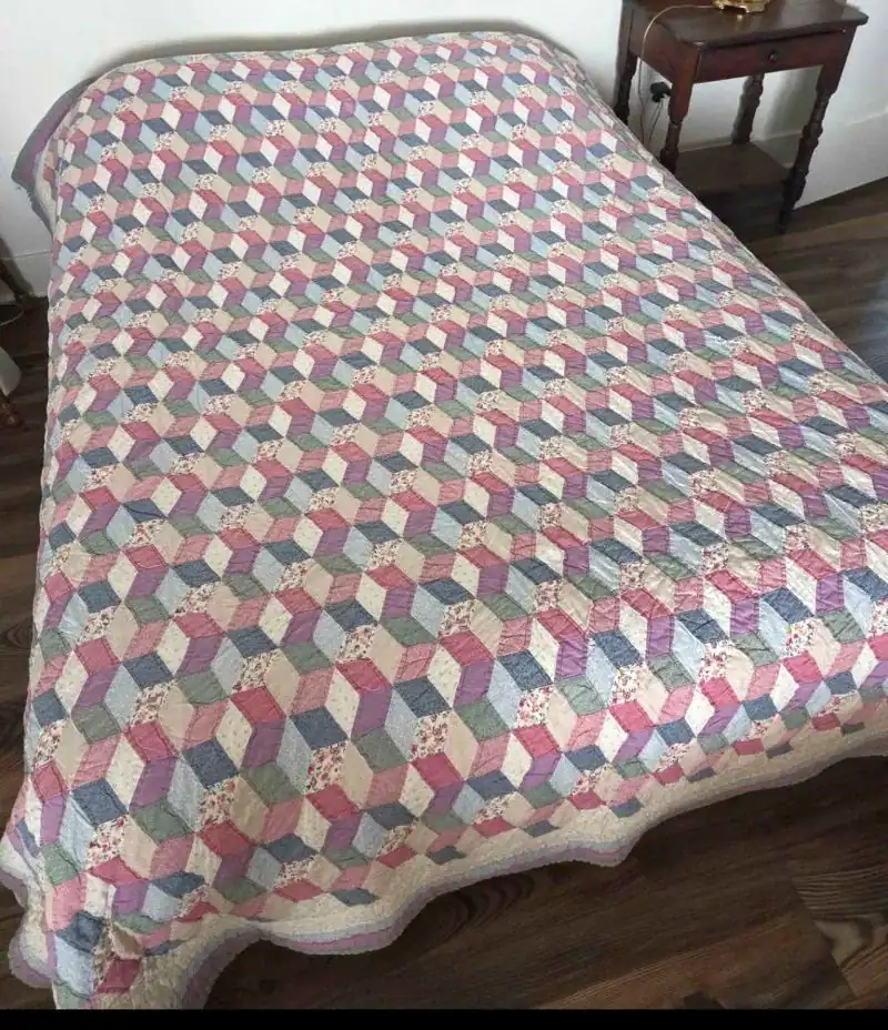 A bed covered with a colorful quilt featuring a geometric cube pattern in pink, blue, cream, and floral prints. A small wooden side table is next to the bed, on a wooden floor.