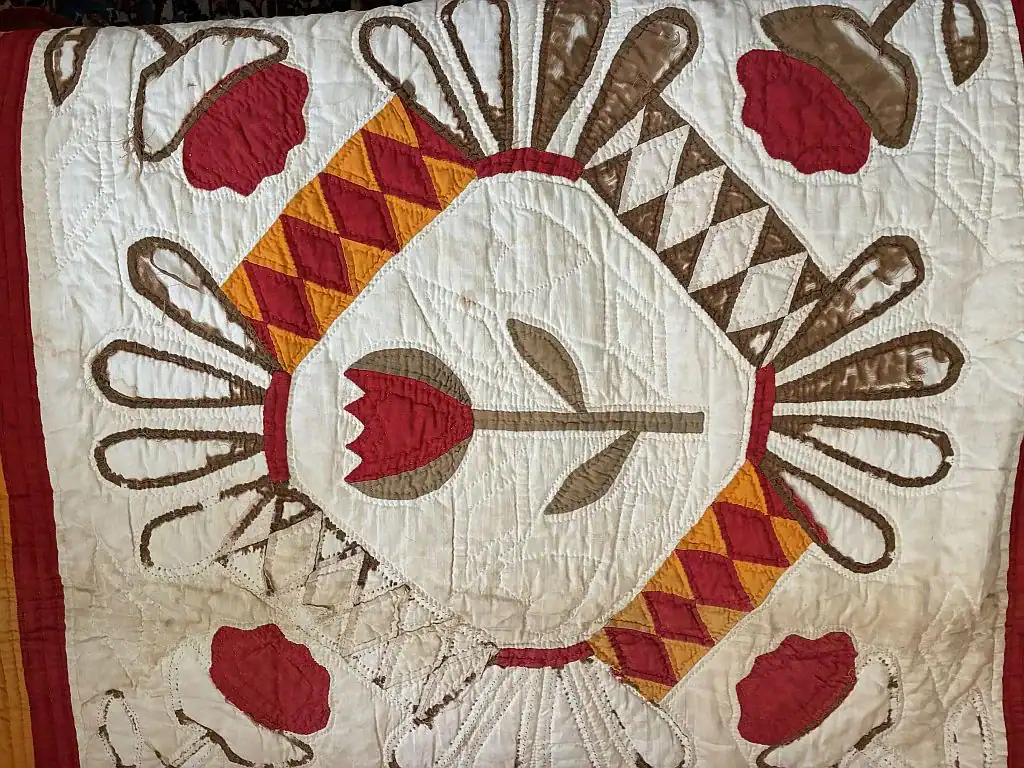A handmade quilt featuring a central tulip design, red and brown flower motifs, and diamond shapes in yellow, brown, and red. The quilt has a symmetrical layout with intricate stitching on a cream background.