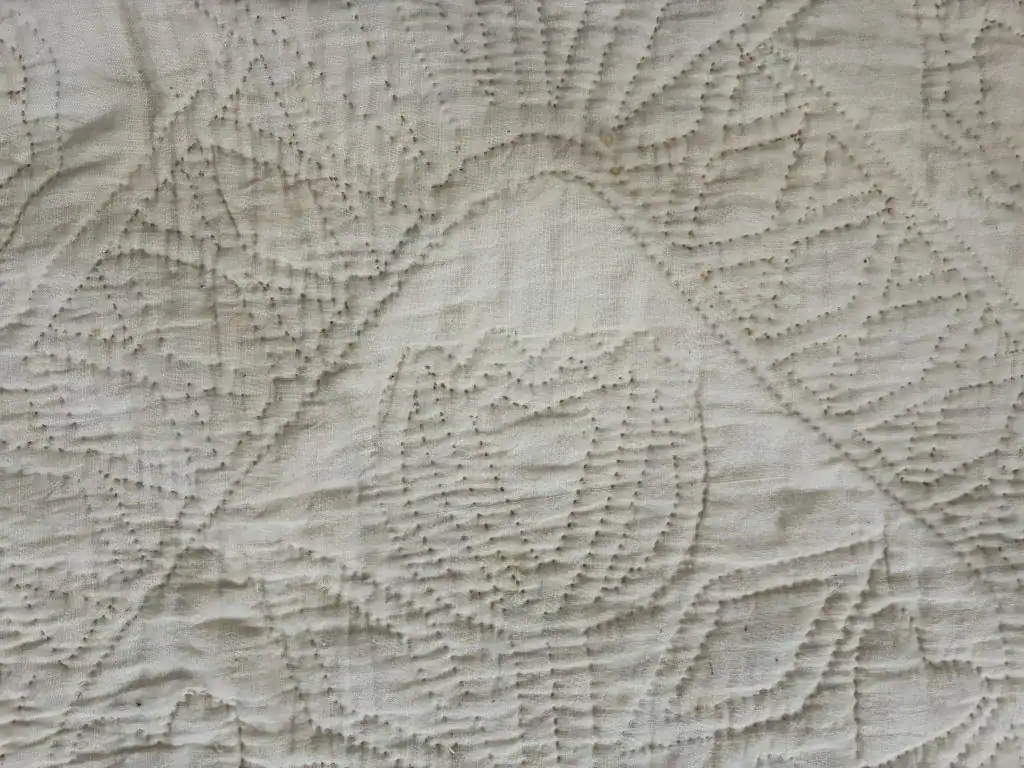 Close-up of a cream-colored quilt with a textured, hand-stitched floral and geometric pattern, featuring raised stitches forming tulip-like shapes and diamonds on the fabric surface.