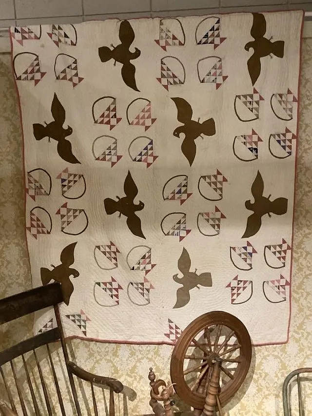 A quilt hangs on a wall, featuring brown bird silhouettes and geometric fan patterns crafted from 1876 Centennial Fabric. A spinning wheel and rocking chair are in front, creating a cozy, vintage scene.