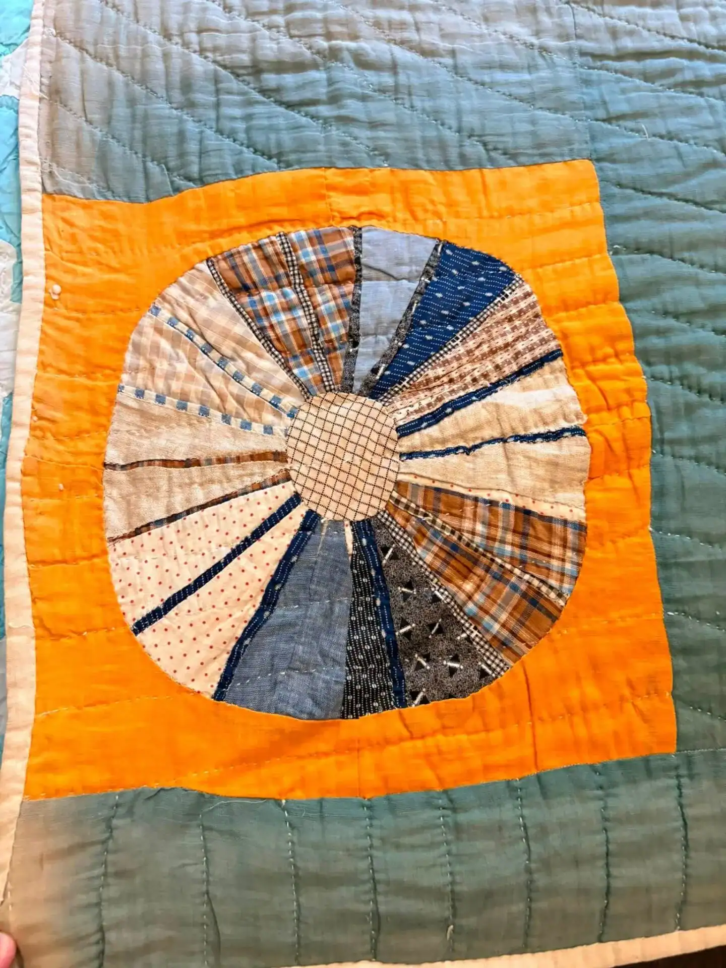 A quilt block with a circular, pie-like patchwork of blue, cream, orange, and patterned fabrics on an orange square, surrounded by light blue quilted fabric.