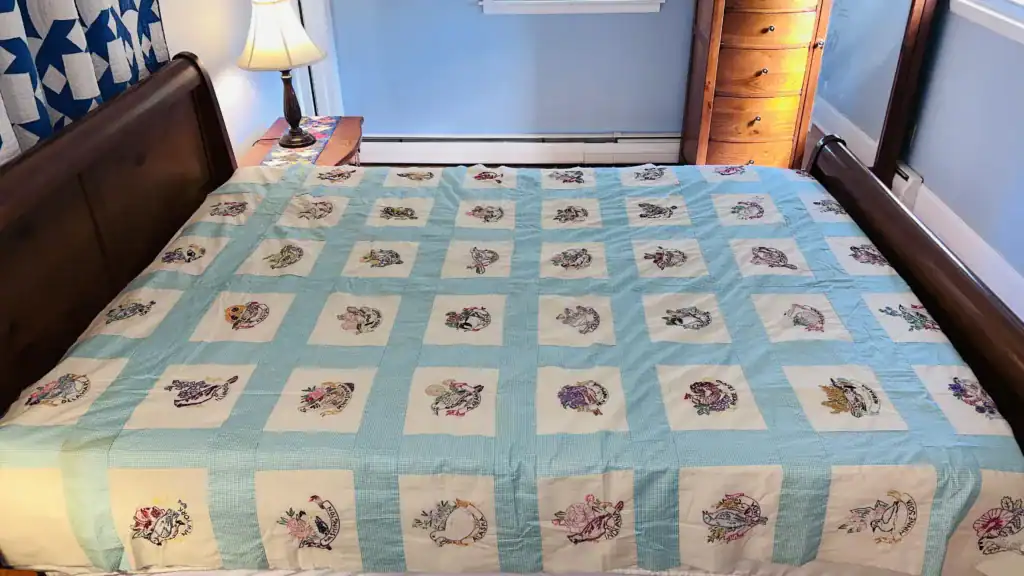 Embroidered-birds-on-bed A wooden bed with a colorful patchwork quilt featuring embroidered flower squares, including State Birds and Flowers Outline Embroidery, next to a nightstand with a lamp, in a blue bedroom with a window and wooden dresser.