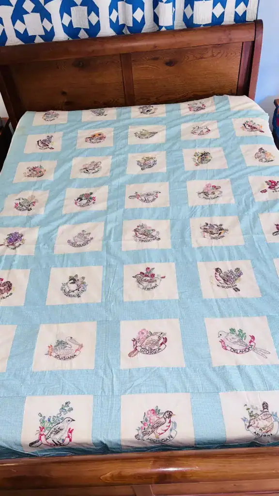 A quilt with blue and white squares, each square featuring colorful floral designs—some inspired by State Birds and Flowers Outline Embroidery—and names of months embroidered in script, spread neatly on a wooden bed.