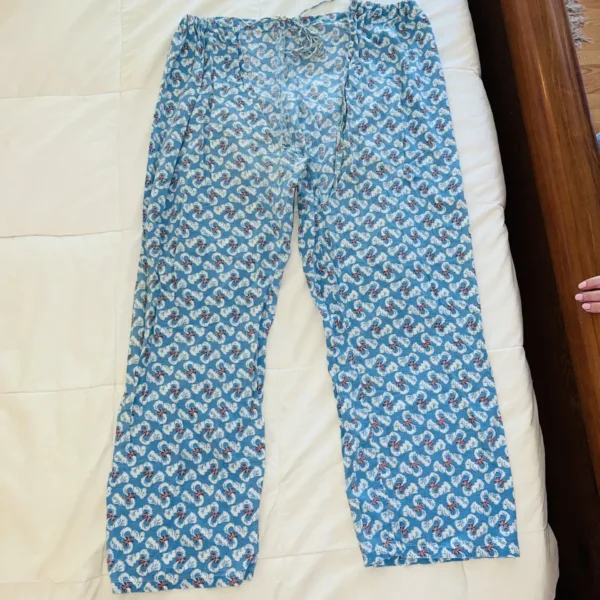 Blue pajama pants with a repeating white seahorse pattern lie flat on a white bedspread next to a wooden bed frame, with part of a hand visible at the edge.