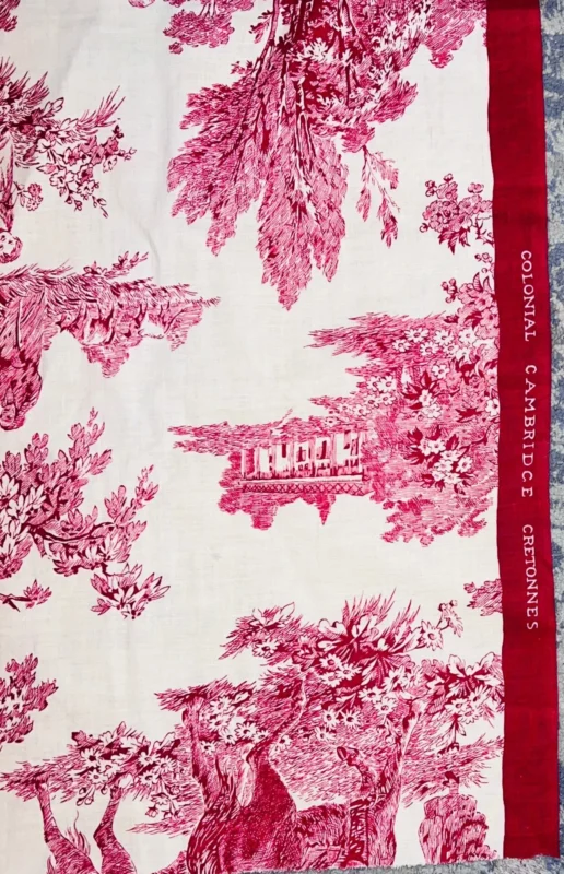 A patterned fabric featuring red pastoral scenes and trees on white. The border is red with white text reading COLONIAL CAMBRIDGE CERDONNES. The edge of the fabric is visible against a blue background.