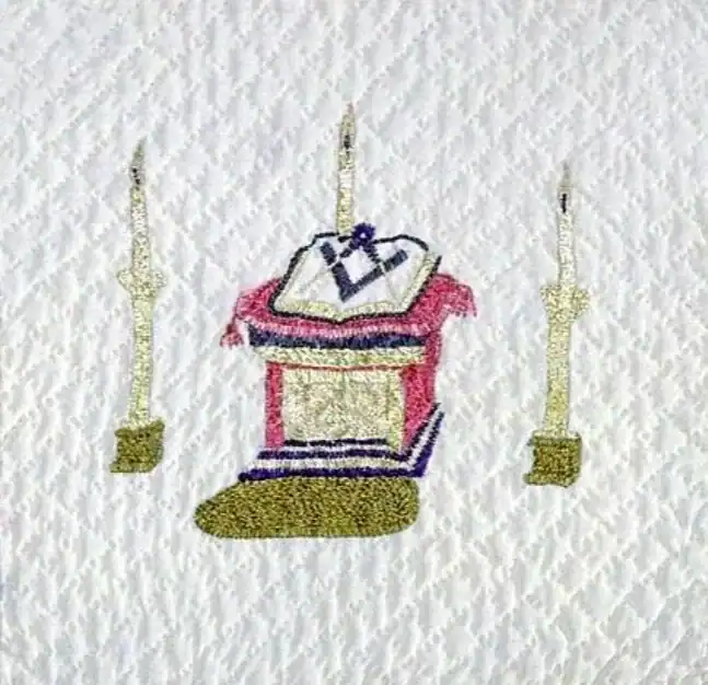 Embroidered image of an altar with an open book and two upright candles or columns on each side, all on a textured white fabric background.