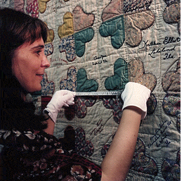 Pensacola-quilt A woman wearing white gloves measures a patchwork quilt decorated with signatures using a measuring tape.