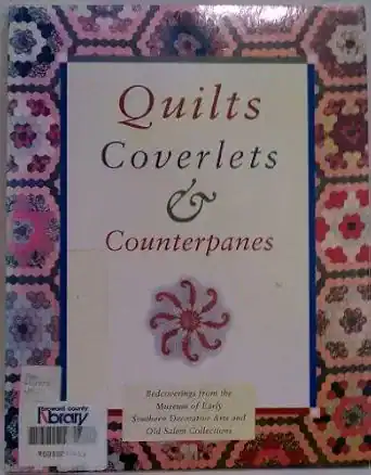 Book cover titled Quilts, Coverlets & Counterpanes with a pink and red hexagon quilt pattern border and a decorative floral motif at the center bottom. Library stickers are visible on the lower left.