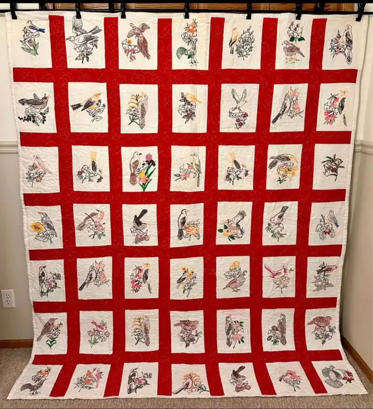 A large quilt with a red grid pattern and white squares, each square featuring colorful embroidered birds and flowers, hangs against a beige wall—a beautiful piece preserved from the March 1997 QHL Archives.