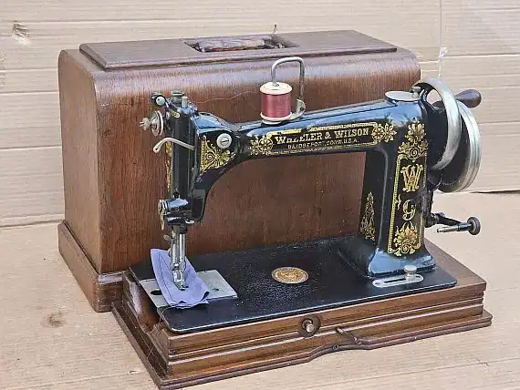 Wheeler-and-Wilson-sewing-machine Antique black and gold sewing machine on a wooden base, with a spool of red thread and an ornate wooden carrying case behind it, set against a cardboard background.