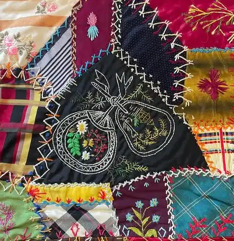 Colorful quilt with various patterned fabric patches, decorative stitching, and detailed white embroidery of two bags filled with flowers at the center. Embroidered floral designs accent some patches.