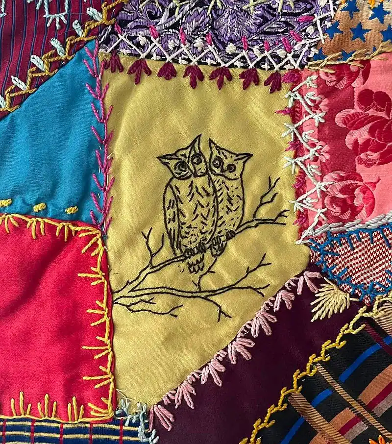 A colorful patchwork quilt features embroidered stitches and two black-stitched owls perched on a branch in the center yellow section, surrounded by bright fabric pieces and decorative borders.