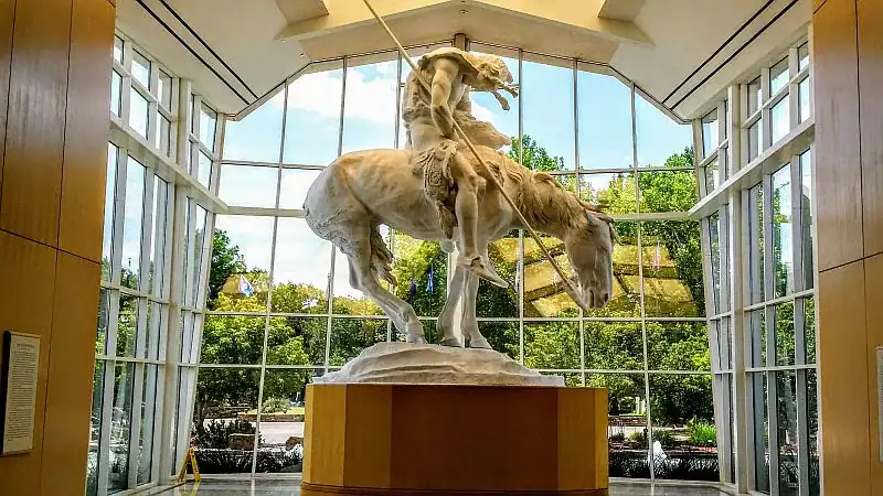 end-of-the-trail A large white statue of a Native American on a horse stands in a sunlit room with tall windows and a view of green trees outside. The figure is slumped forward, holding a spear.