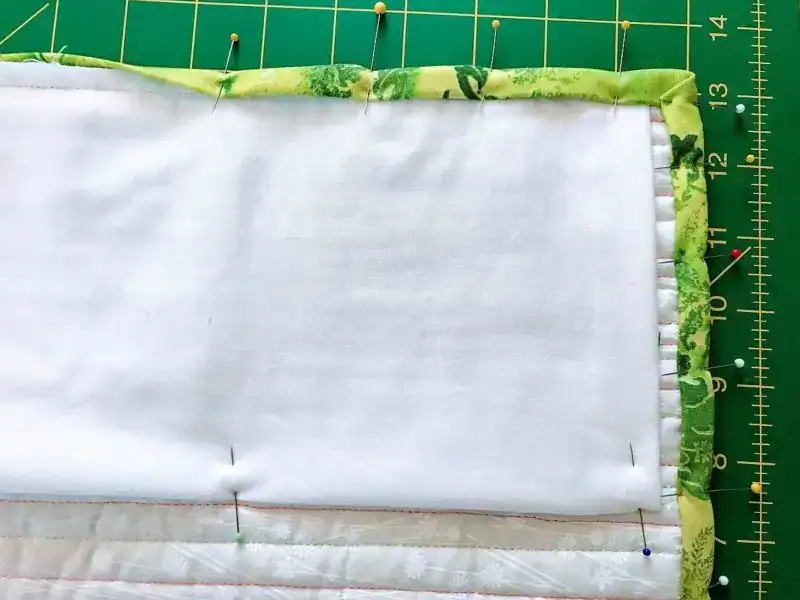 Hanging Sleeve A quilt with white fabric and green floral binding is pinned along the edge. It is placed on a green cutting mat with a yellow measurement grid.