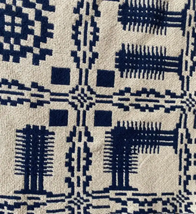 Blue geometric, grid-like patterns and stylized shapes are woven into beige fabric, resembling traditional textile or quilt designs. The stitching forms symmetrical lines, squares, and abstract tree-like motifs.