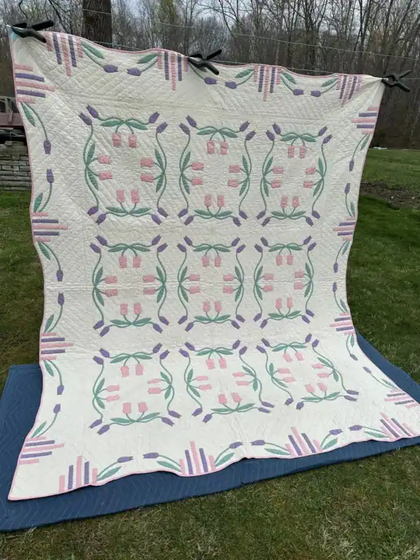 A large handmade quilt with a floral and geometric pattern in pink, green, and purple on a white background hangs outdoors on a clothesline. Trees and grass are visible in the background. This beautiful piece was featured in the March 1997 QHL Archives.