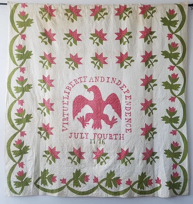 A quilt crafted with 1876 Centennial Fabric features a striking red eagle in the center, encircled by the words VIRTUE, LIBERTY AND INDEPENDENCE, JULY FOURTH 1776, and beautifully adorned with pink and green floral patterns.