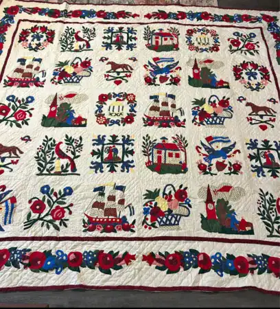 A cream-colored quilt featuring a grid of appliquéd squares decorated with folk art motifs including ships, houses, birds, and floral baskets, surrounded by a floral border.